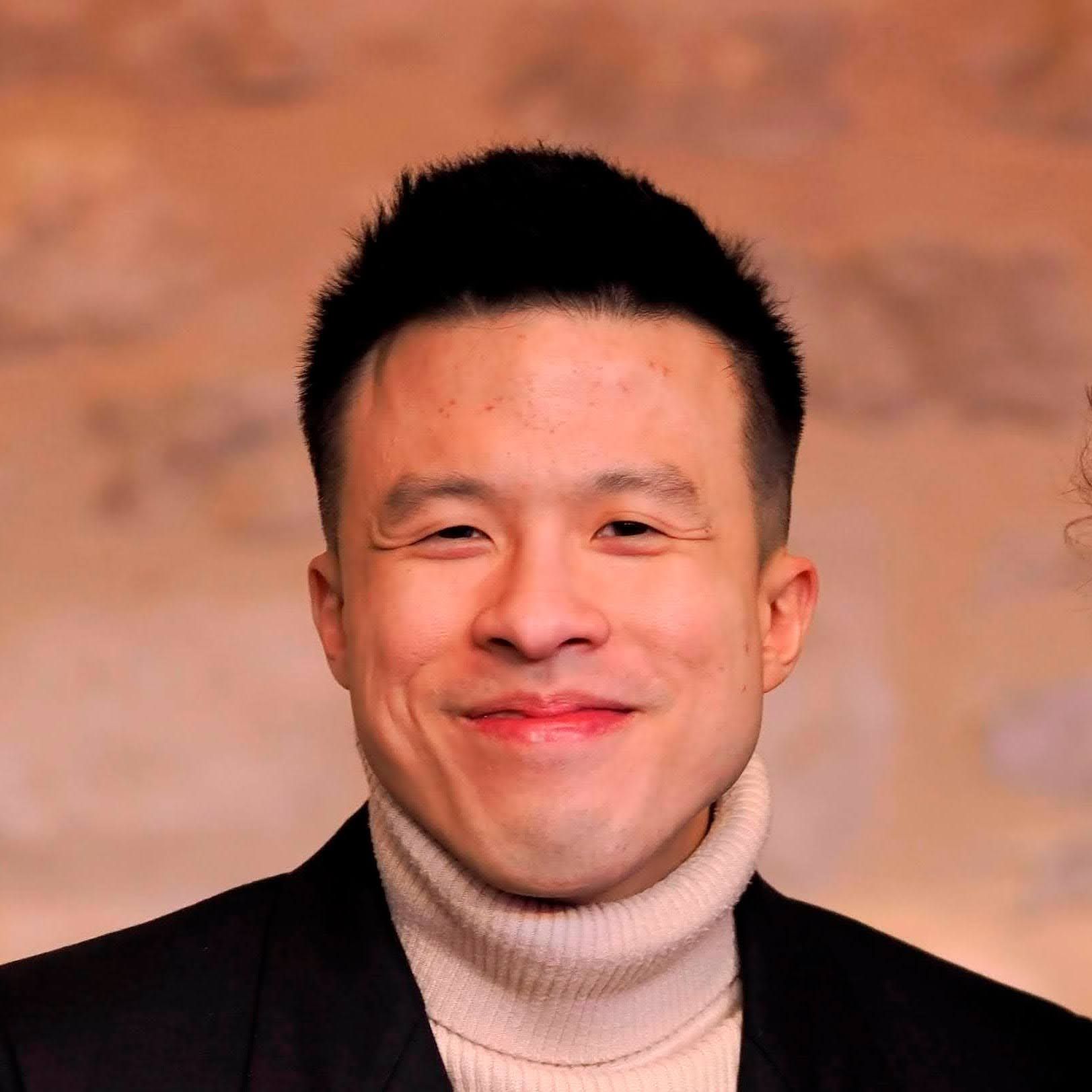 Jason, in a black jacket and white turtleneck sweater smiles warmly at the camera.