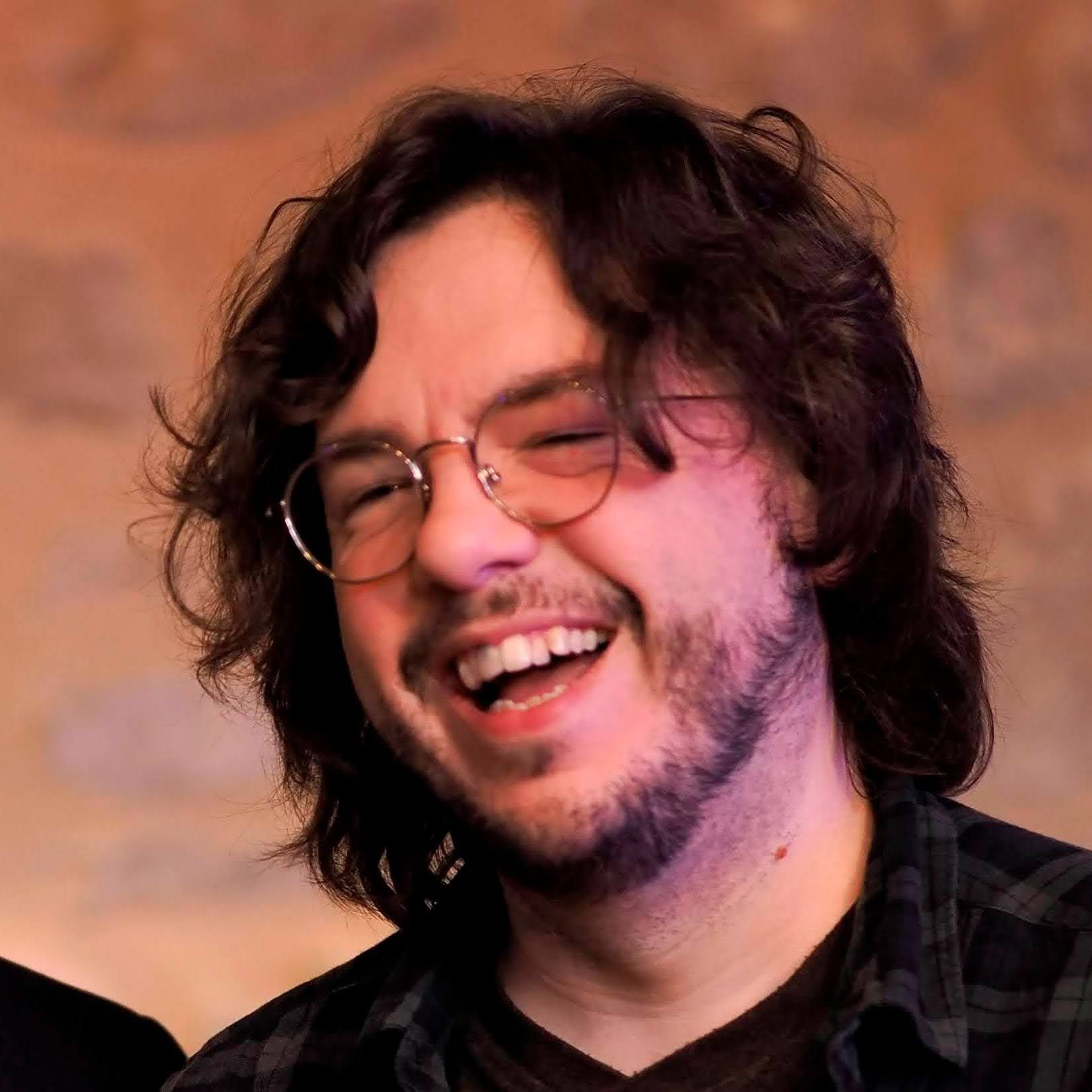 Tom, with shaggy brown hair, round glasses, and short beard, laughs at something off-camera.